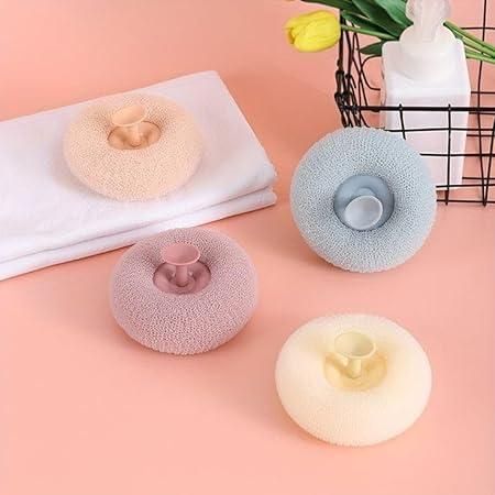 Four donut-shaped bath sponges in different colors on a pink surface with a towel and flowers in the background.