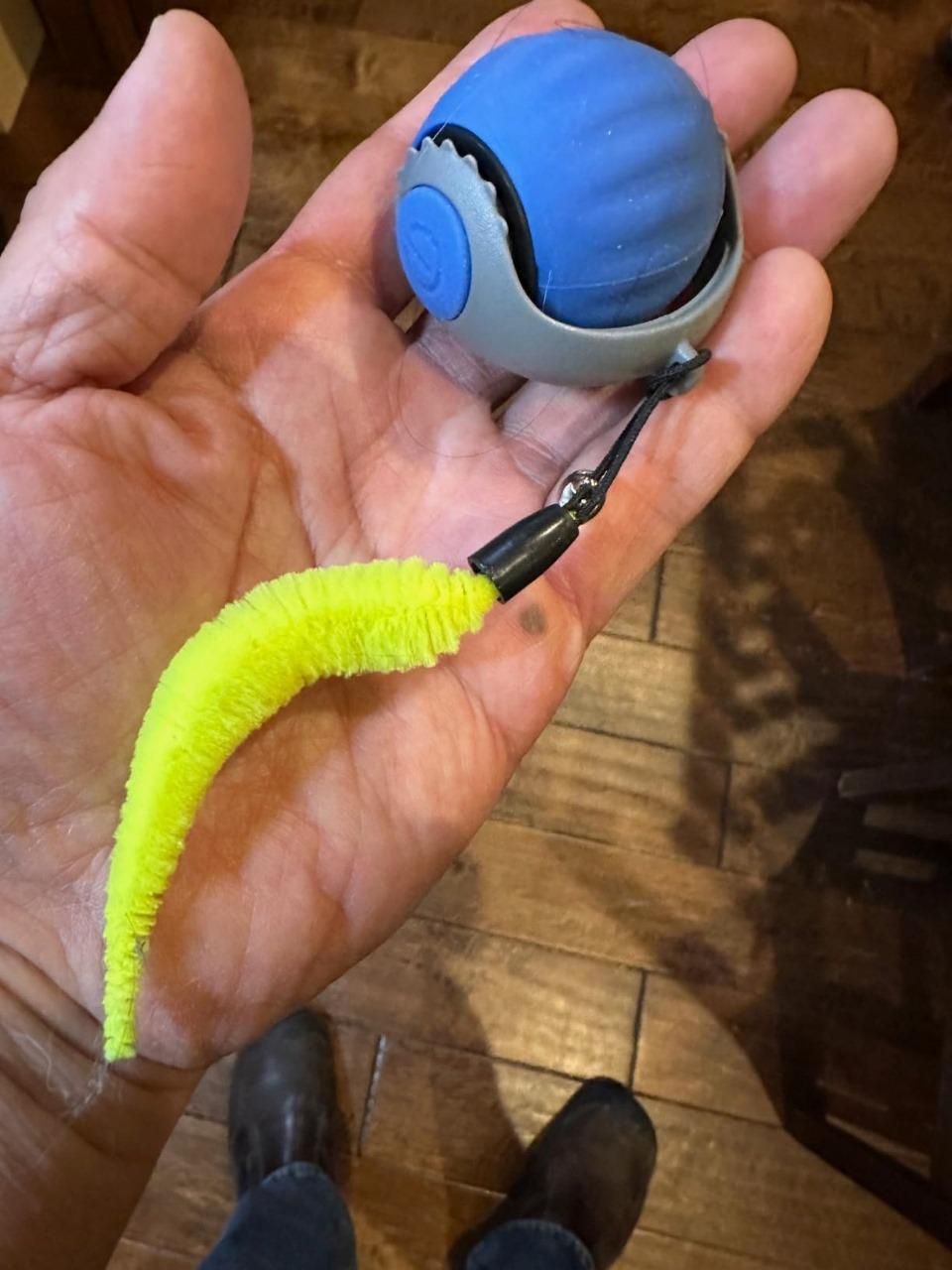 Hand holding a blue and gray ball with a yellow handle on a wooden floor.