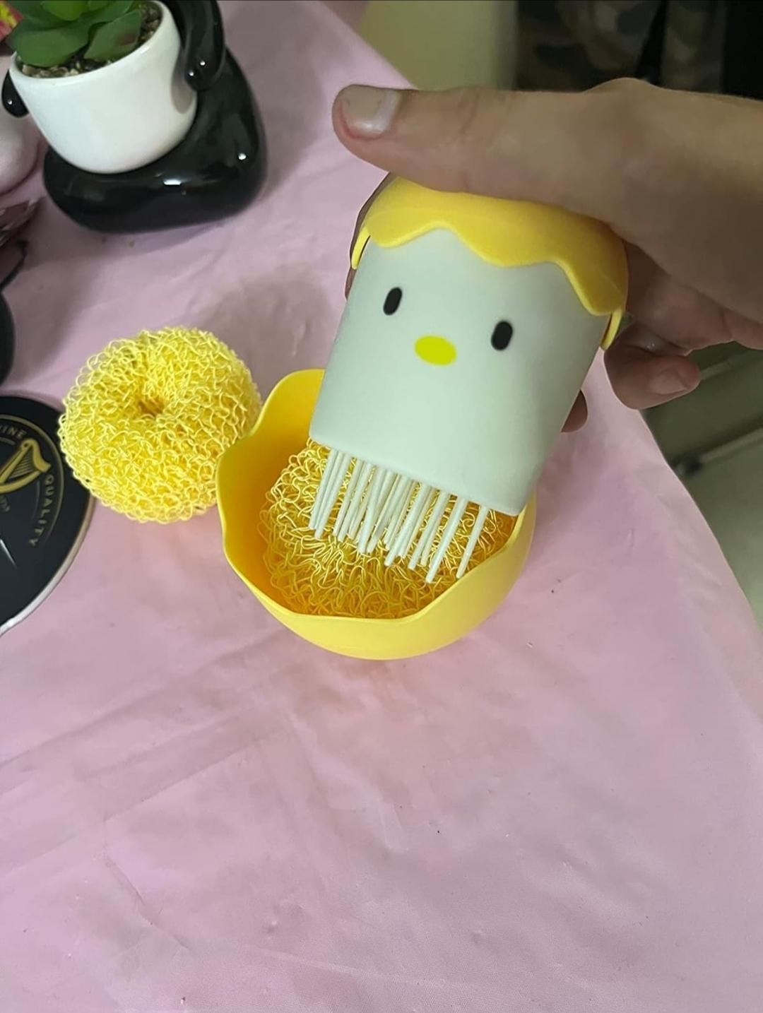 Hand holding a yellow and white cleaning brush with a cartoon face on a pink surface.