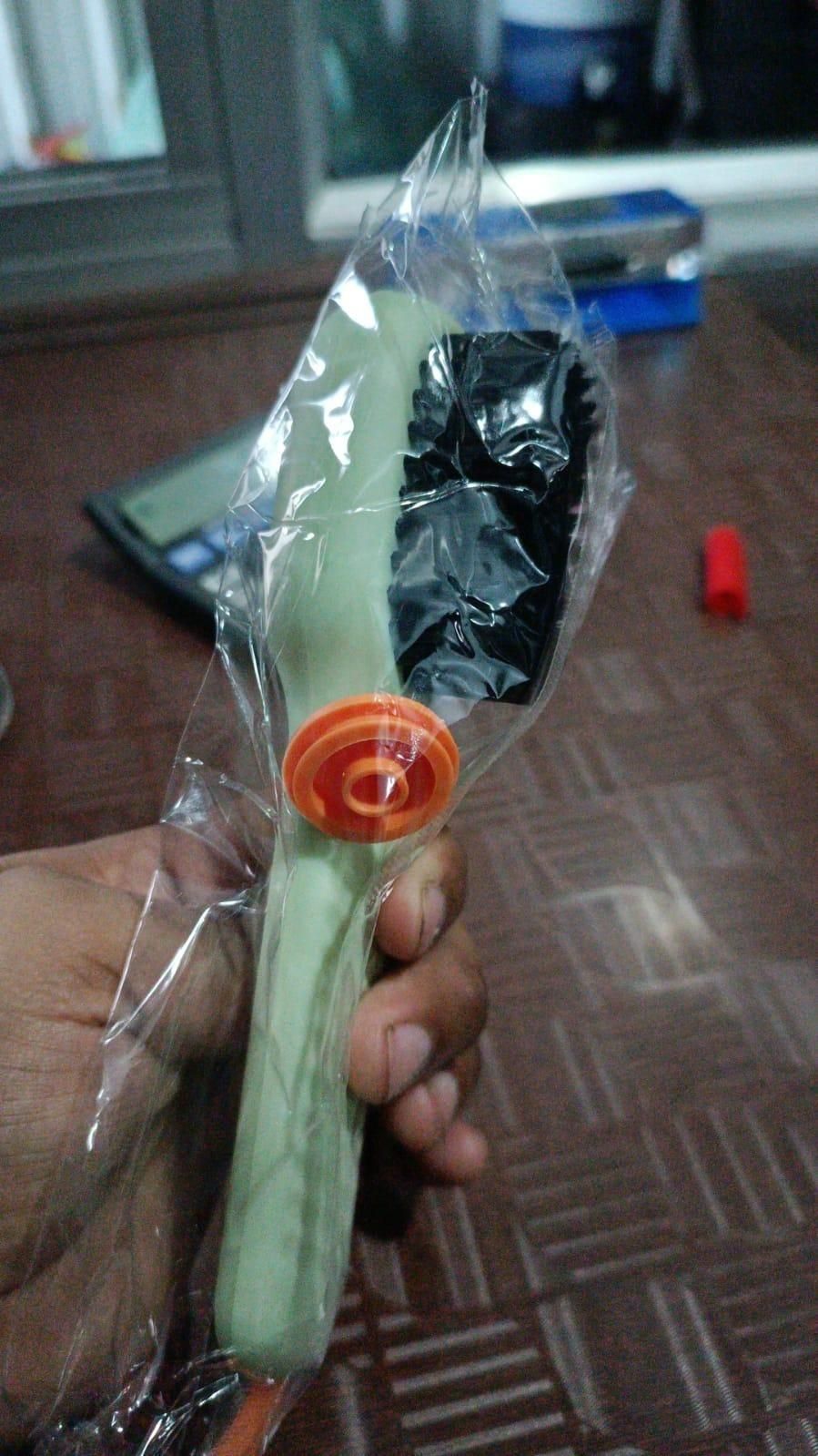 Hand holding a green object with an orange handle wrapped in clear plastic, on a carpeted floor.
