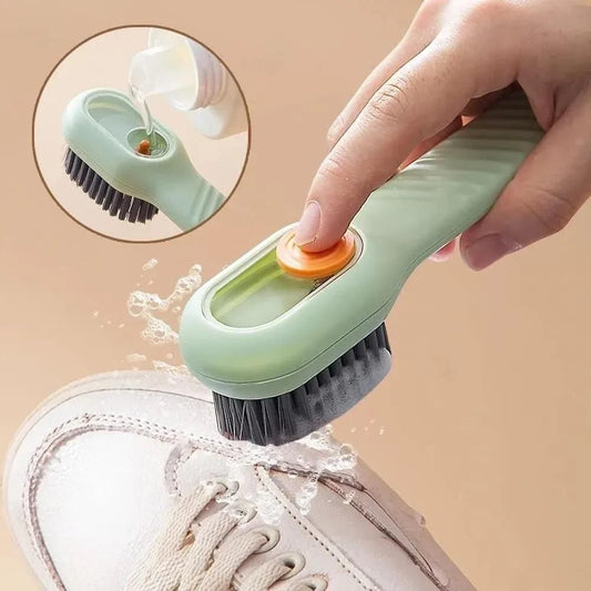 Hand using a green cleaning brush with an orange button to scrub a white sneaker on a beige background.